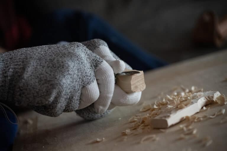 A closeup of a person carving wood with a knife while wearing gloves